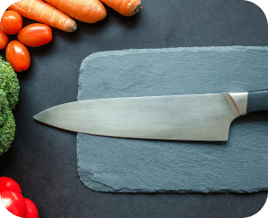 A flat lay of a chef's knife on a black cutting board with fresh vegetables surrounding it.